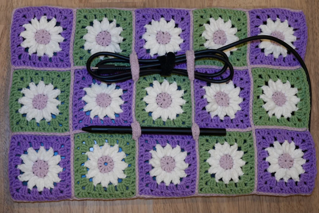 Top view of crocheted table sleeve with cable and pen attached by 4 little loops crocheted onto the joining seam in pink yarn. One of the cable's ends is plugged into the tablet within the sleeve which the rest sits on top of the sleeve in a folded coil constrained by the new loops