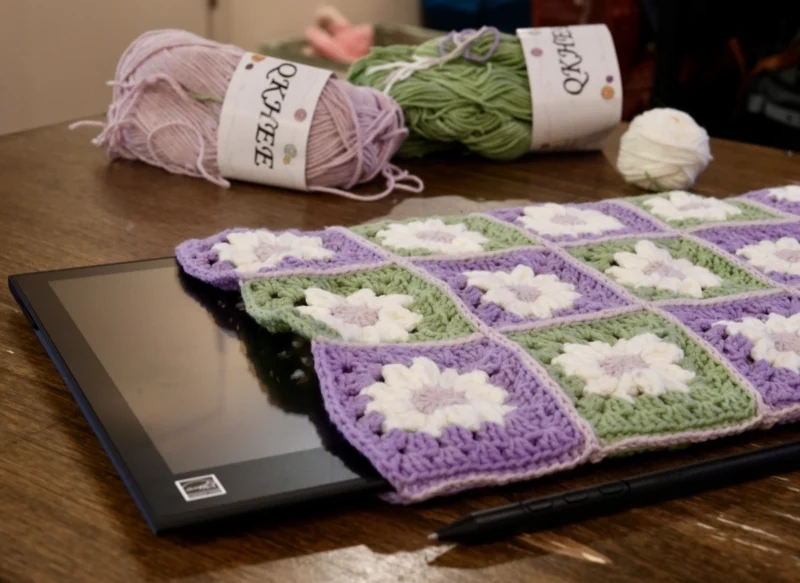 Wacom Movink drawing tablet peeking out of a custom made tablet sleeve made of crocheted granny squares with white daisies and alternating green and purple backgrounds.