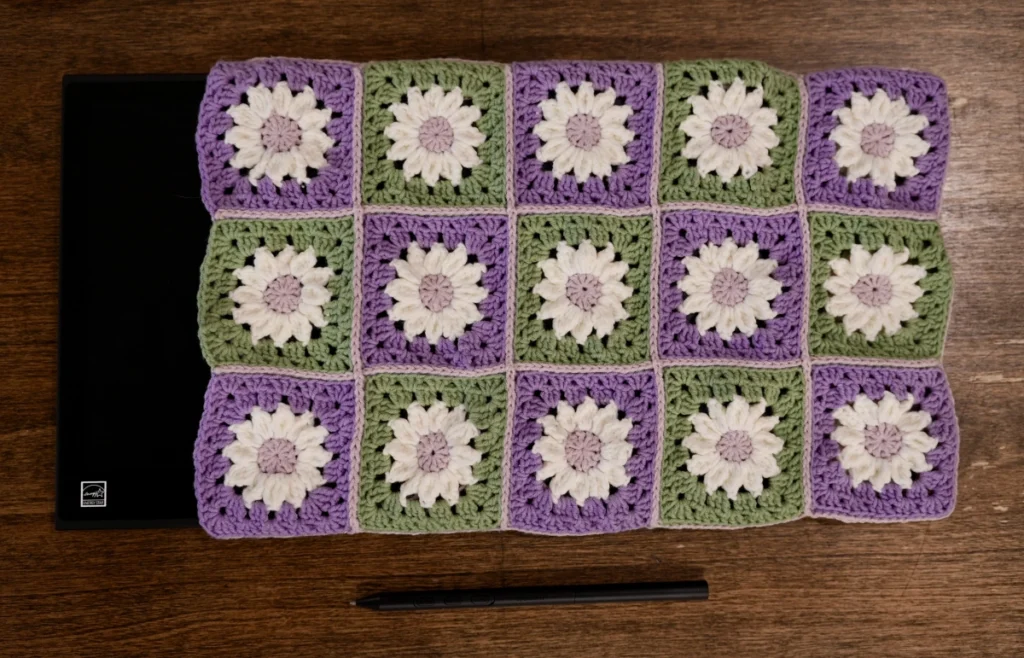 The front of a made tablet sleeve for a Wacom Movink made of 15 granny squares with white daisies in the middle and alternating green and purple backgrounds. Each square is joined using pink yarn in a slightly raised slip stitch. 