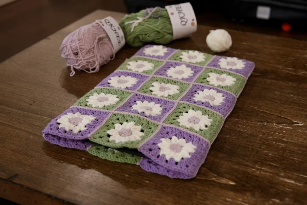 Tablet sleeve made of 30 joined granny squares, 15 on each side. The squares have a daisy pattern in white with pink centers and backgrounds in alternating green and purple. They are sewn together with pink yarn. 2 yarn skeins in pink and green sit beside the tablet sleeve, along with a small ball of white yarn. 